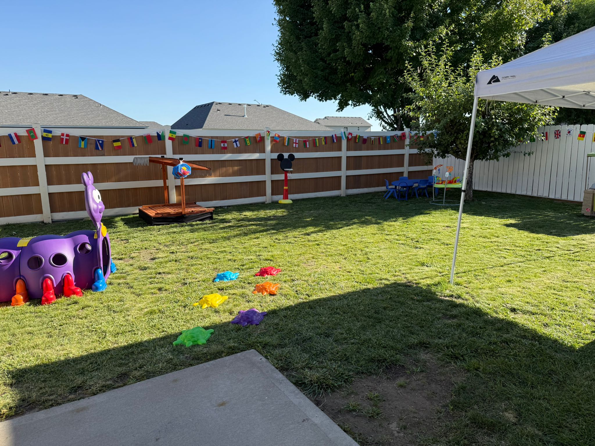 Patio trasero con una cerca blanca, equipo de juego colorido, un área cubierta, mesas y banderas internacionales colgadas en la cerca.