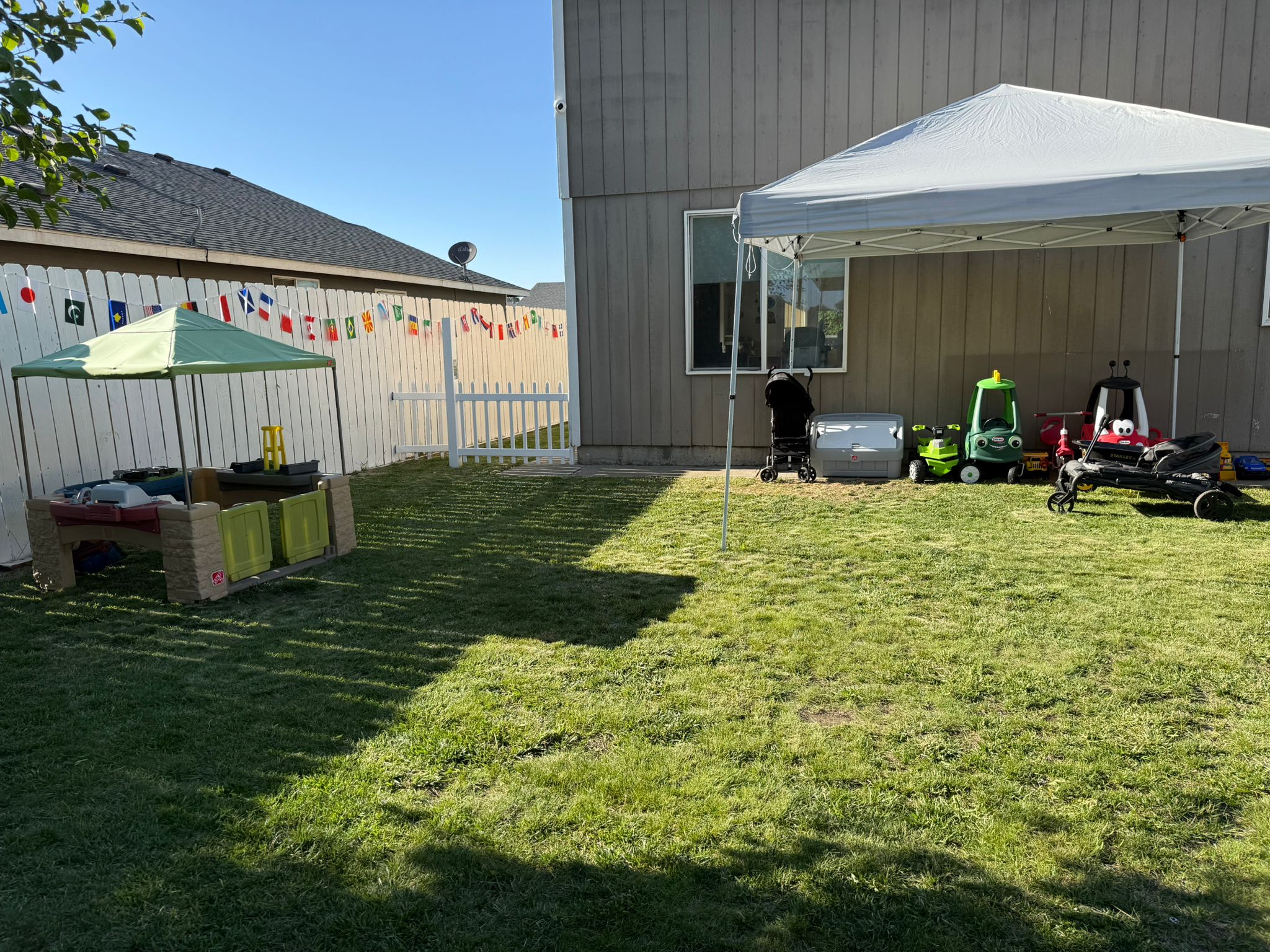 Patio trasero con un toldo, una casita de juegos para niños, vehículos de juguete y una nevera portátil sobre el césped, junto a una casa con cerca blanca decorada con banderines.