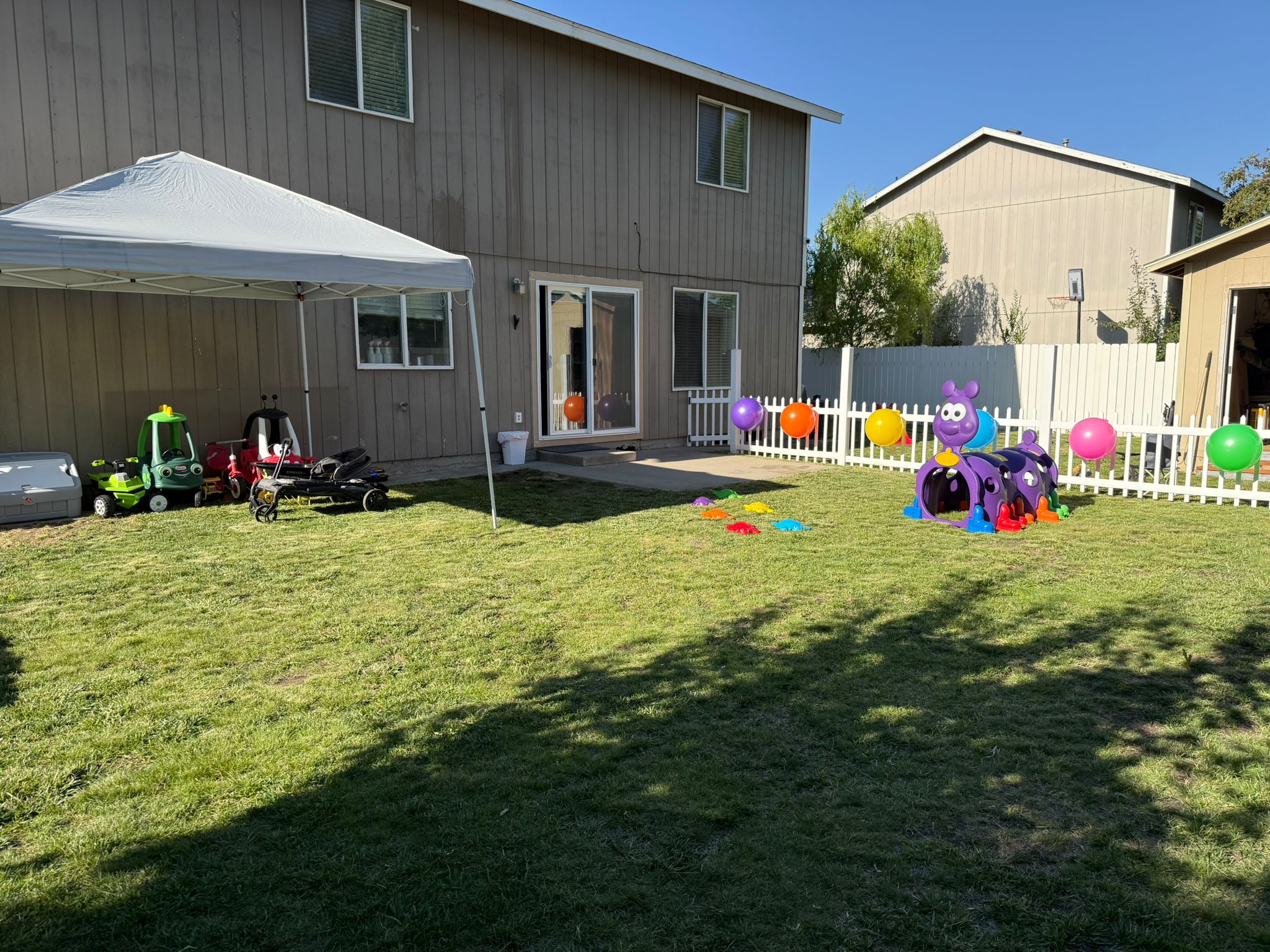 Patio trasero con césped, una cerca blanca con globos de colores, un toldo con carritos de juguete y un túnel de oruga morado.
