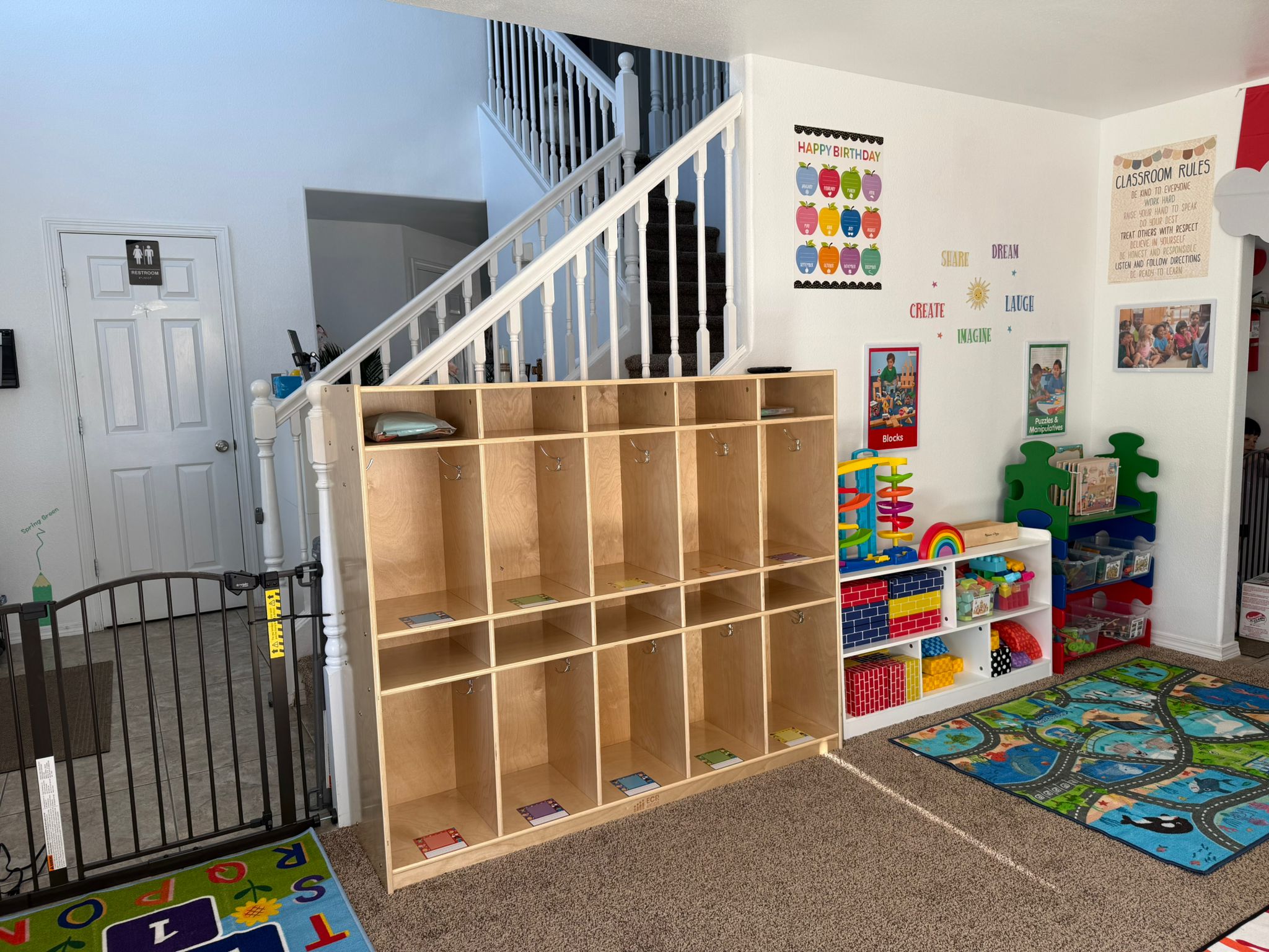 Aula ordenada con casilleros de madera, juguetes de colores, estanterías, una alfombra de juegos con carreteras y carteles educativos en las paredes.