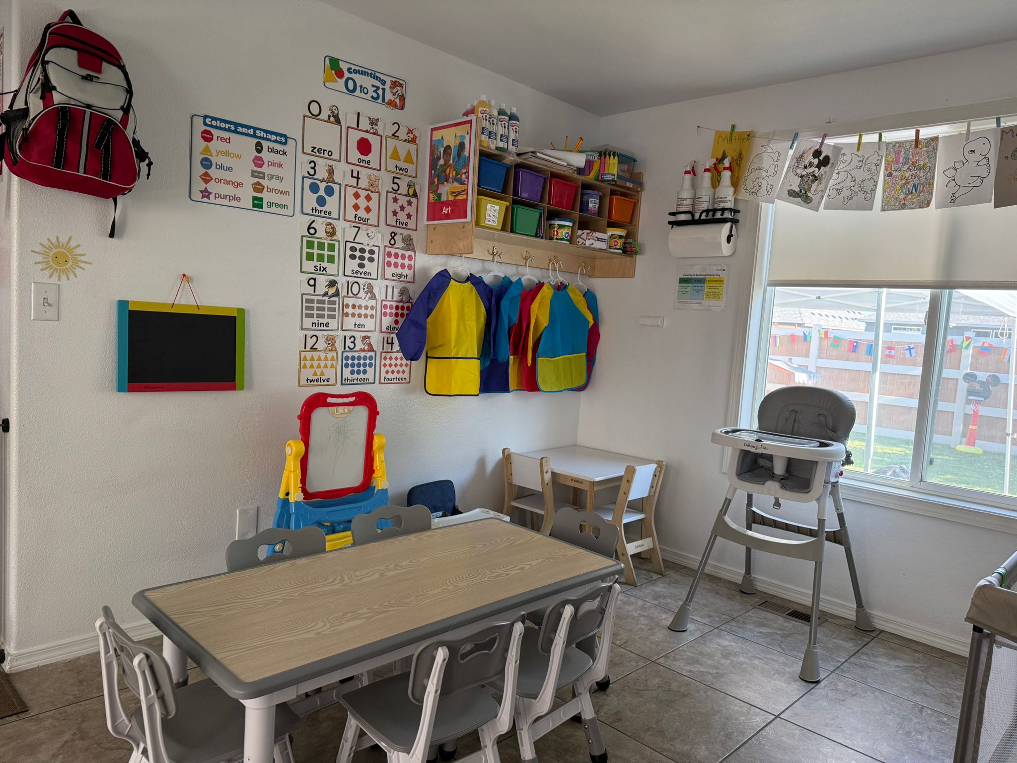 Aula de preescolar colorida con mesas pequeñas, sillas, materiales de arte, carteles educativos, disfraces y obras de arte de los niños colgadas en la ventana.