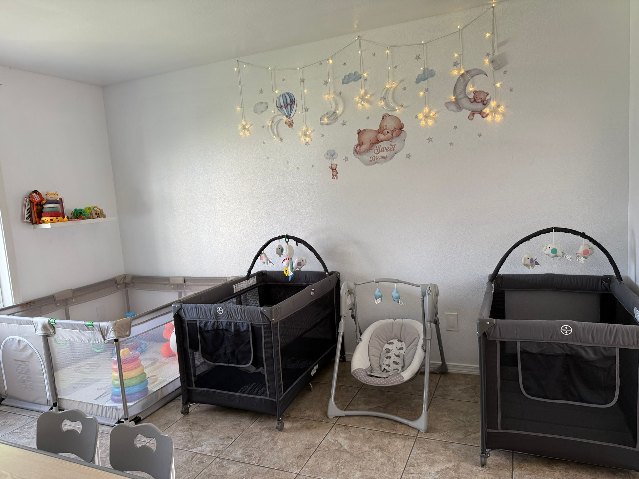 Sala de cunas con tres corrales, un columpio para bebé y juguetes. La pared está decorada con nubes, estrellas, globos aerostáticos y un letrero de Dulces Sueños.