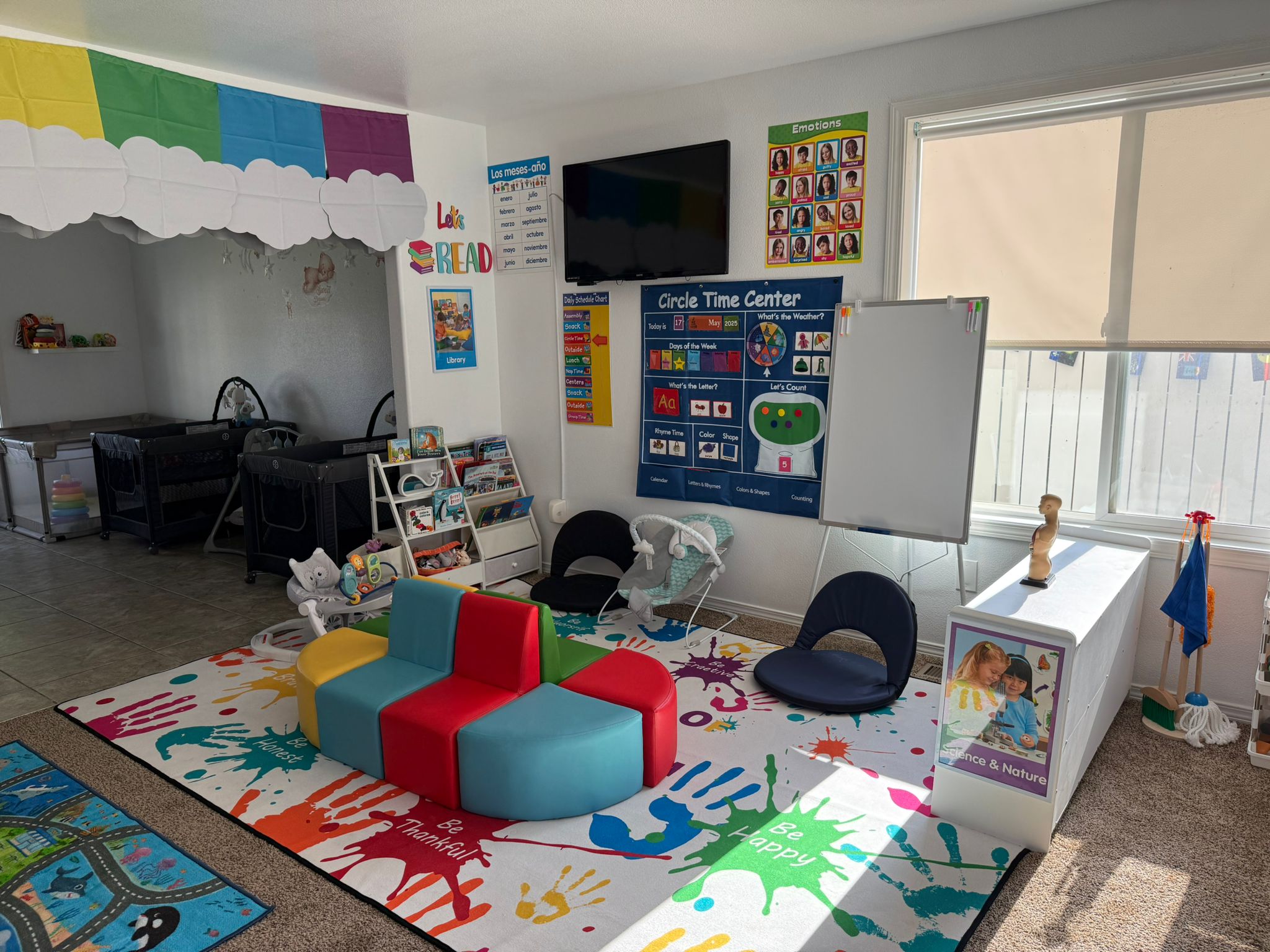 Aula luminosa con asientos de colores, carteles educativos, estanterías, un televisor y una alfombra con huellas de manos.