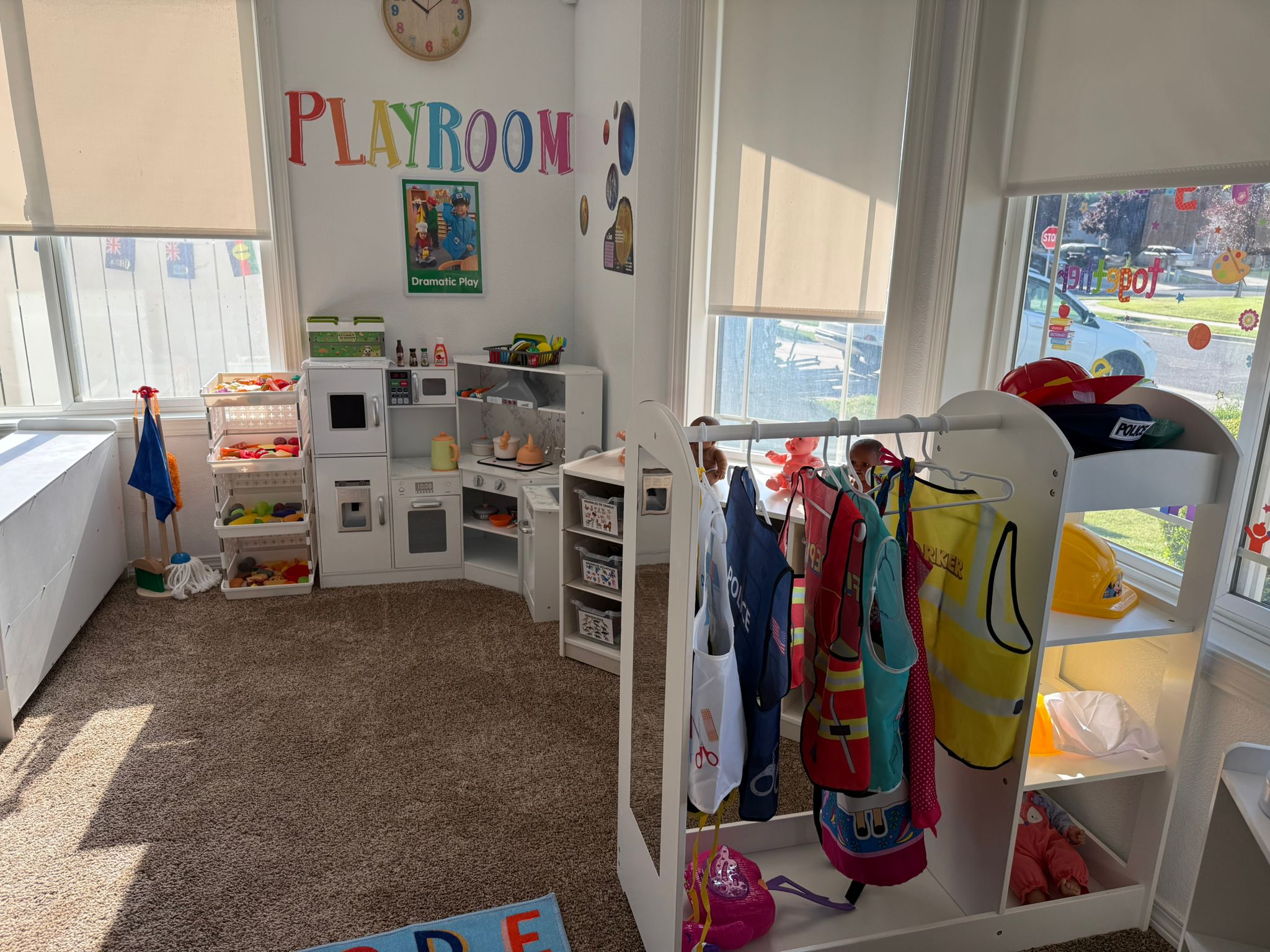 Sala de juegos luminosa con una cocinita, disfraces, comida de juguete, contenedores de almacenamiento y la palabra PLAYROOM en la pared.